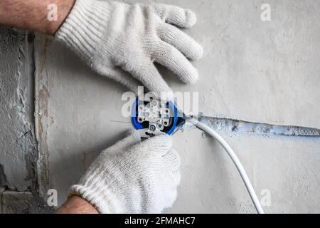 Nahaufnahme von den Händen eines kaukasischen Elektroinstallatrikers Eine weiße Steckdose an einer grauen Wand eines House.Home Reparaturen während der Pandemie und Aufenthalt Stockfoto