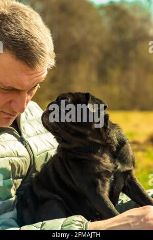 Der Besitzer des Rüden zwinkert seinem süßen schwarzen Moorhund zu Sitzt auf seinem Schoß auf dem natürlichen Boden des Frühlingsparks Stockfoto