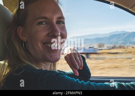 Lächelnde Frau schaut auf die Kamera, während sie auf der Autoreise fährt In der Wüste Stockfoto