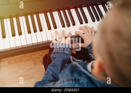 Kleiner Junge, der zu Hause E-Piano spielt, die Sonne trifft auf die Tastatur. Er trägt eine Jeansjacke, seine blonden Haare in einem Brötchen. Verschwommen, hoher Winkel, Nahaufnahme Stockfoto