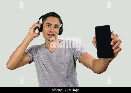 Der 28-jährige Brasilianer mit einem überraschten Gesicht, mit einer Hand auf dem kabellosen Kopfhörer, zeigt das Smartphone der Kamera. Stockfoto
