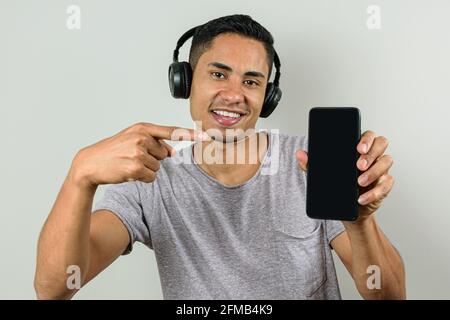 Lächelnder, 28-jähriger Brasilianer mit kabellosem Kopfhörer, zeigt mit dem Finger auf sein Smartphone und zeigt es der Kamera. Stockfoto