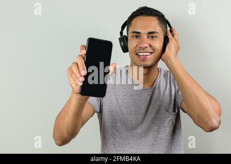 Lächelnder 28-jähriger Brasilianer mit einer Hand auf dem kabellosen Kopfhörer und der anderen zeigt sein Smartphone der Kamera. Stockfoto