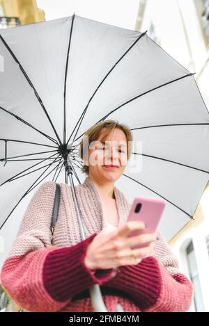 Vertikales Porträt einer blonden kaukasischen Frau, die ihr Telefon benutzt. Sie ist auf der Straße und hält einen Regenschirm Stockfoto