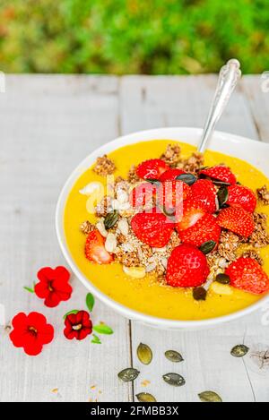 Fruchtiger Frühstücks-Smoothie aus pürierter Mango mit Erdbeeren und Müsli auf weißem Holz im Garten. Stockfoto