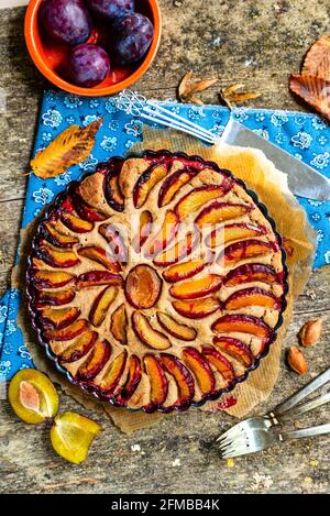 Pflaumenkuchen auf rustikalem Holzhintergrund, blaublühendes Tuch, geschnittene Pflaumen, Herbstblätter, Tonschüssel mit frischen Pflaumen. Stockfoto