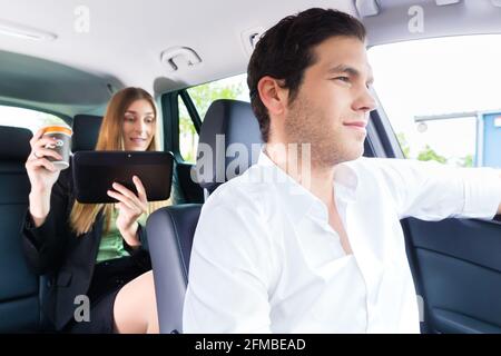 Junge Taxifahrer fahren Geschäftsfrau im Taxi, sie halten Tasse Kaffee und einem Tablet-computer Stockfoto