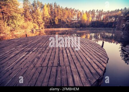 Magischer Sonnenaufgang über dem See. Pinien am Seeufer. Ruhiger See am frühen Morgen. Hölzerner runder Pier am See. Naturlandschaft. Stockfoto