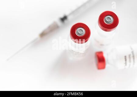 Dünne Spritze neben einigen Impffläschchen mit roter Kappe auf weißem Hintergrund. Konzentrieren Sie sich auf die rote Kappe auf der linken roten Flasche, beabsichtigte Unschärfe Stockfoto