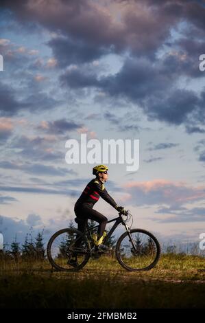 Vertikale Momentaufnahme eines Radfahrers bei Sonnenuntergang in den Bergen. Fahrradfahrer trägt Helm, Sportbrille und Uniform. Seitenansicht. Abendhimmel im Hintergrund. Konzept des aktiven Lebensstils Stockfoto