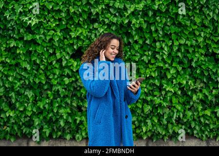 Junge lateinerin mit ihrem Smartphone und ihrem Kopfhörer am Straße mit grünem Hintergrund Stockfoto