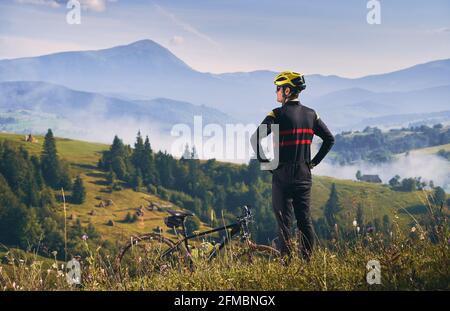 Rückansicht eines Radfahrers in Uniform und Helm, der auf einem Hügel steht und die wunderschöne Berglandschaft beobachtet. Am frühen Morgen in den neblig sonnigen Bergen. Konzept der Einheit mit der Natur Stockfoto