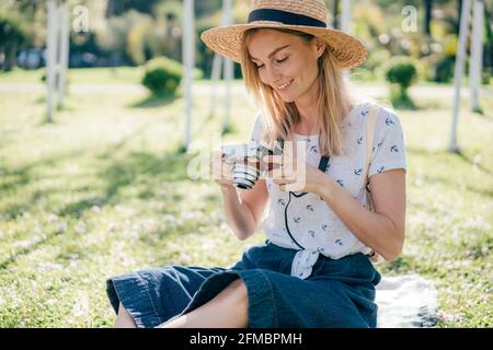 Ein Mädchen in einem Strohhut sitzt auf dem Gras im Park und schaut auf ein Foto auf einer Digitalkamera. Professioneller Fotograf. Frau auf einer Sommerreise. Stockfoto