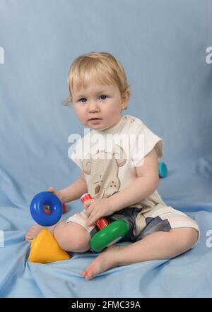 Ein kleines blondes Kind mit blauen Augen sitzt auf einem blauen Hintergrund und spielt mit Spielzeug. Mehrfarbige Kunststoffpyramide Stockfoto