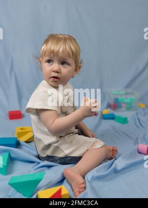 Ein kleines blondes Mädchen in einem kurzen Jumpsuit sitzt und Spielt mit bunten Würfeln mit einem Konstrukteur Stockfoto