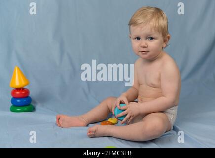 Ein kleines blondes Mädchen in einer Windel sitzt auf einem blauen Hintergrund und spielt mit einer hölzernen farbigen Pyramide. Blick auf die Kamera Stockfoto