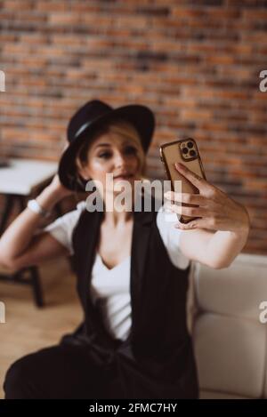 Attraktive stilvolle blonde Frau in schwarzem Hut lächelnd macht Selfie auf ihrem Smartphone.Weiche selektive Fokus, Unschärfe, Kunst Lärm. Stockfoto