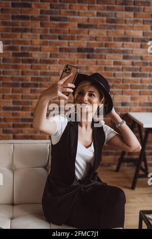 Attraktive stilvolle blonde Frau in schwarzem Hut lächelnd macht Selfie auf ihrem Smartphone.Weiche selektive Fokus, Unschärfe, Kunst Lärm. Stockfoto