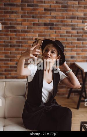 Attraktive stilvolle blonde Frau in schwarzem Hut lächelnd macht Selfie auf ihrem Smartphone.Weiche selektive Fokus, Unschärfe, Kunst Lärm. Stockfoto