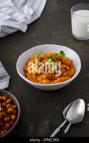 Dicke Kürbissuppe mit gebackenen Kichererbsen und Rettichsprossen auf einem Teller mit einem Glas Milch auf dem Tisch, leckeres gesundes Abendessen Stockfoto