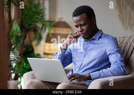 Junger ernsthafter afrikanischer Mann, der zu Hause auf dem Laptop tippt Stockfoto