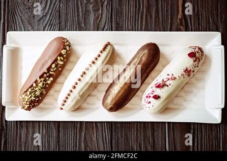 Französische Dessert-Eclare oder Profiterolen mit Schokoladenglasur, mit verschiedenen Belägen auf einem weißen Teller, Holzhintergrund. Gebäck Pudding Kuchen mit cr Stockfoto