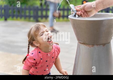 Das kleine Mädchen trinkt fröhlich Wasser aus einem Trinkbrunnen Der Park an einem Sommertag Stockfoto