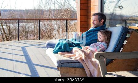 Vater und Tochter sitzen auf einem Stuhl auf der Terrasse unter einer Wolldecke zusammen. Familienurlaub außerhalb der Stadt Konzept. Vater und Tochter sind d Stockfoto