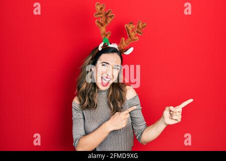 Junge hispanische Mädchen mit Hirsch weihnachtshut lächelnd und Blick auf die Kamera mit zwei Händen und Fingern zur Seite zeigen. Stockfoto