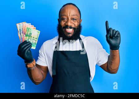 Junger afroamerikanischer Mann mit professioneller Schürze, der Südafrika hält rand Banknoten lächeln mit einer Idee oder Frage, die mit dem Finger zeigt Mit glücklich Stockfoto