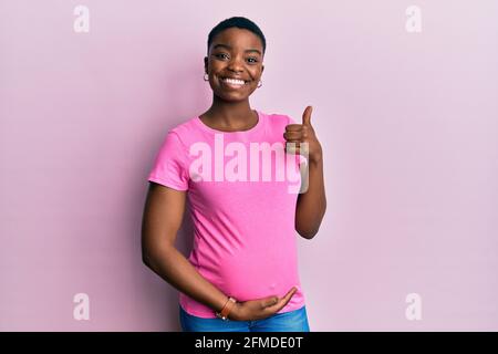 Junge afroamerikanische Frau erwartet ein Baby, berühren schwanger Bauch tun glückliche Daumen nach oben Geste mit der Hand. Genehmigung Ausdruck Blick auf die Ca Stockfoto