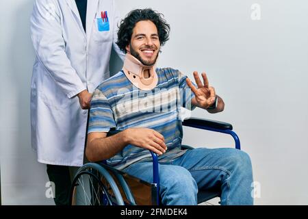 Gutaussehender hispanischer Mann, der auf dem Rollstuhl sitzt und einen Halsband trägt und mit den Fingern Nr. drei nach oben zeigt, während er selbstbewusst und glücklich lächelt. Stockfoto