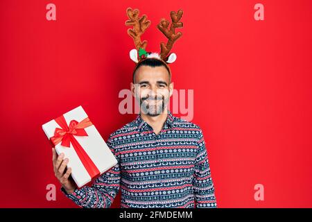 Junger hispanischer Mann trägt Hirsch weihnachtshut hält Geschenk suchen Positiv und glücklich stehen und lächeln mit einem selbstbewussten Lächeln Zeigt Zähne Stockfoto