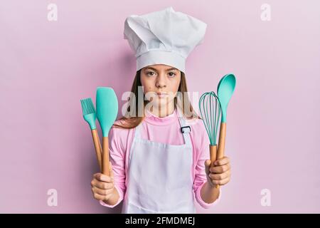 Schöne Brünette kleines Mädchen trägt professionelle Koch Schürze hält Kochwerkzeuge entspannt mit ernsten Ausdruck auf Gesicht. Einfach und natürlich looki Stockfoto