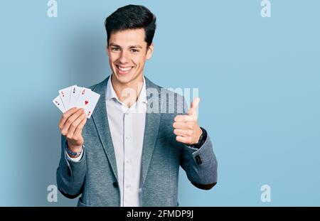 Junger hispanischer Geschäftsmann, der Poker spielt und Karten hält, die glücklich und positiv lächeln, Daumen nach oben, indem er ausgezeichnete und Zustimmungszeichen macht Stockfoto