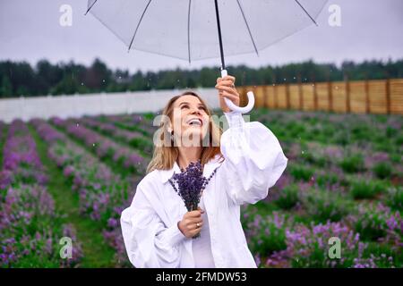 Fröhliche Frau tanzt unter Regen, hält Regenschirm und Blumenstrauß und lächelt im bewölkten Himmel. Foto mit geringer Tiefenschärfe Stockfoto