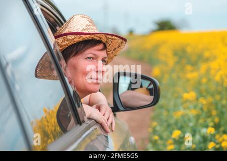 Frau genießt die Autofahrt in blühender Sommerlandschaft, Kopf mit Strohhut, der aus dem Fenster reicht, selektiver Fokus Stockfoto