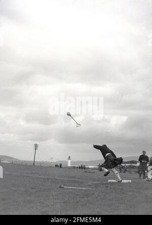 1956, historisch, draußen auf einem Grasfeld bei einem Hochlandspiel, ein männlicher Konkurrent in einem Kilt, beim Hammerwurf, Schottland, Großbritannien. Der Hammer, der geworfen wird, besteht aus einer kugelförmigen Metallkugel, die an einem Griff befestigt ist, mit der der Hammer um ihren Kopf geschwirbelt und dann so weit wie möglich geworfen wird, wobei die größte Entfernung der Gewinner ist. In den Highland-Spielen gibt es keine Seitenlinie, im Gegensatz zum Hammerwurf bei den Olympischen Spielen. Stockfoto