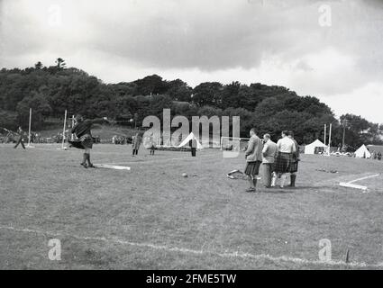 1956, historisch, draußen auf einem Grasfeld bei einem Hochlandspiel, ein männlicher Konkurrent in einem Kilt, der am Hammerwerfen-Wettbewerb teilnimmt, beobachtet von Beamten, Schottland, Großbritannien. Der Hammer besteht aus einer Kugelkugel aus Metall, die an einem Griff befestigt ist, mit dem der Hammer um ihren Kopf geschwirbelt und dann so weit wie möglich geworfen wird, wobei die größte Entfernung der Gewinner ist. In den Highland-Spielen gibt es keine Seitenlinie, im Gegensatz zum Hammerwurf bei den Olympischen Spielen. Stockfoto