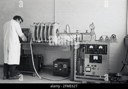 1963, historisch gesehen, ein weiß beschichteter männlicher Techniker, der neben Geräten steht, die von einem Oszilloskop getestet wurden, oft einfach als Scope oder O-Scope bekannt, eine Art elektronisches Testinstrument, das Signalspannungen grafisch anzeigt und Spannungswellen misst. Stockfoto