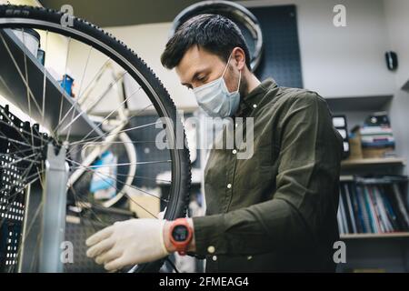 Fahrradmechaniker trägt Schutzmaske und Handschuhe repariert Kunden Fahrrad Rad in Übereinstimmung mit Quarantänestandards während des Coronavirus Stockfoto