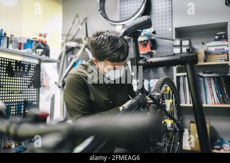 Mechaniker repariert Fahrrad in der Werkstatt trägt covid 19 Maske. Arbeiter des Fahrradspeichers fixiert Fahrrad während der Coronavirus-Quarantäne in Schutzmaske Stockfoto