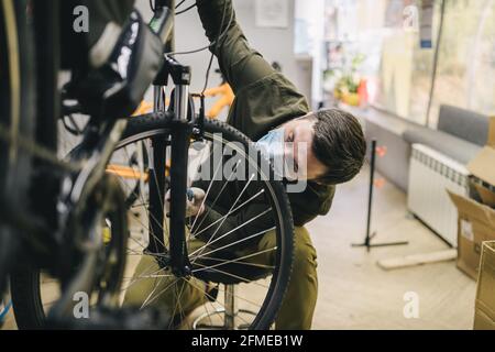 Fahrradmechaniker trägt Schutzmaske und Handschuhe repariert Kunden Fahrrad Rad in Übereinstimmung mit Quarantänestandards während des Coronavirus Stockfoto