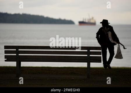 Ein Mann, der einen Cowboyhut trägt und eine Gitarre und Plastiktüte an einer Parkbank vorbeiführt, wird auf seinem Spaziergang entlang der English Bay in Vancouver, Briti, als Silhouette geziert Stockfoto