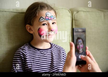 Kind mit Gesichtsbemalung von Kitty, nettes kleines Mädchen mit gemalter Maske auf Gesicht Posen für Foto. Glücklich hübsches Kind mit schönen Make-up als Zeichnung. Ador Stockfoto