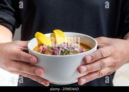 Hände eines Mannes, dessen Gesicht nicht sichtbar ist und einen Teller Encebollado hält, ein typisches Meeresfrüchte-Gericht aus Ecuador mit Fisch, Zwiebeln und Yucca. Stockfoto