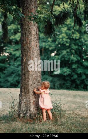 Nettes Baby Mädchen ein Jahr alt in rosa Kleid stehen bei hohen Baum und erkunden Lernen Naturwelt. Blick von hinten. Kleines, winziges Kind, Kind t Stockfoto