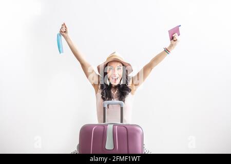 Glückliche weibliche Reisende steht vor ihrem Koffer, bewaffnet sich als Einschränkungen, hält Pass in der einen und Gesichtsmaske in der anderen Hand. Stockfoto