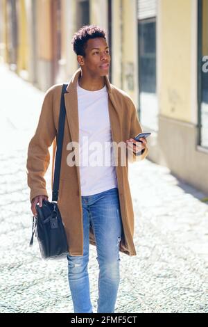 Ein junger schwarzer Mann, der die Straße entlang läuft, trägt eine Aktentasche und ein Smartphone. Stockfoto