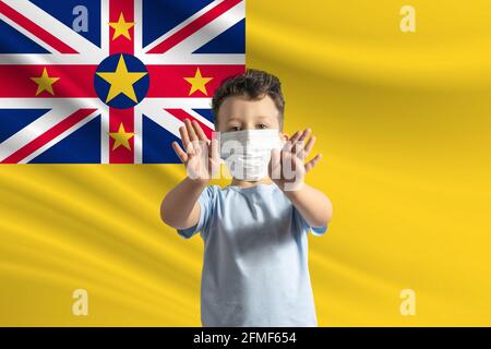 Kleiner weißer Junge in einer Schutzmaske auf dem Hintergrund der Flagge von Niue. Macht mit seinen Händen ein Stoppschild, bleibt zu Hause Niue. Stockfoto